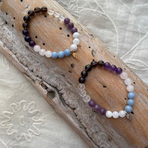 Puede incluir: Dos pulseras hechas con cuentas de piedras preciosas. Las pulseras están superpuestas sobre un trozo de madera a la deriva. La pulsera superior tiene cuentas marrones, transparentes, moradas, blancas y azules. La pulsera inferior tiene cuentas marrones, transparentes, moradas, blancas y azules. Ambas pulseras tienen un dije dorado en forma de ala.