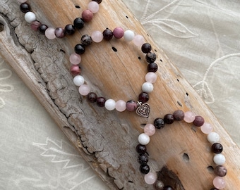 May include: Three bracelets made of pink, white and dark brown beads with a heart charm on each. The bracelets are on a piece of driftwood.