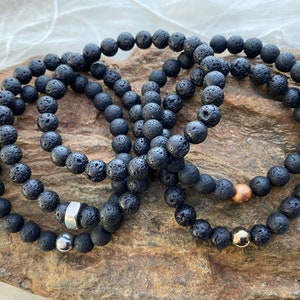 May include: Three black lava rock beaded bracelets with silver and gold accents. The bracelets are stacked on top of each other.