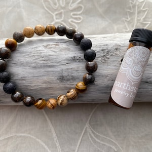 Op de afbeelding: Een bruine, zwarte en gouden kralen armband met een klein flesje etherische olie. De fles heeft een wit etiket met de tekst "earthstone jewelry by".