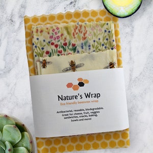 May include: A set of three beeswax food wraps with different patterns. The wraps are yellow with a honeycomb pattern, floral pattern, and a bee pattern. The wraps are packaged together with a white label that reads "Nature's Wrap Eco friendly beeswax wrap".