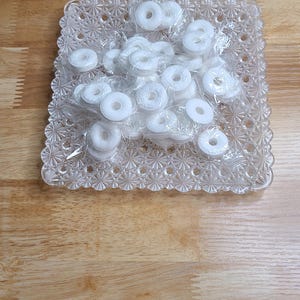 May include: A square, clear glass dish with a textured, star-like pattern on the rim and base. The dish contains a pile of small, white, ring-shaped candies, each individually wrapped in clear plastic film, creating a cluster of white circles.