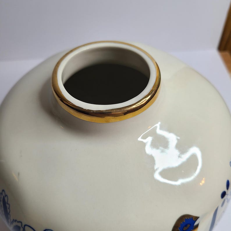 Puede incluir: Un jarr&oacute;n de cer&aacute;mica color crema con un borde dorado. El jarr&oacute;n tiene forma redonda y est&aacute; decorado con motivos florales azules. El interior del jarr&oacute;n es oscuro. El borde dorado a&ntilde;ade un toque de elegancia al dise&ntilde;o.