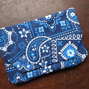 May include: A small blue and white bandana-patterned pouch with a folded flap. The pouch is made of fabric and has a paisley design.