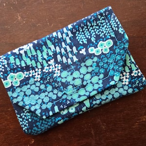 May include: A small, rectangular wallet with a flap closure. The wallet is covered in a dark blue fabric with a pattern of teal and white floral and abstract shapes. The stitching is teal.