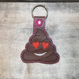 May include: A brown leather keychain shaped like a smiling poop emoji with heart eyes. The keychain has a pink stitched border and a pink button on the strap. A silver key ring is attached.