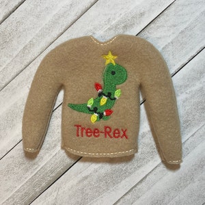 May include: A brown elf-sized sweater with a green dinosaur wearing Christmas lights and a star on top. The text "Tree-Rex" is embroidered below the dinosaur.