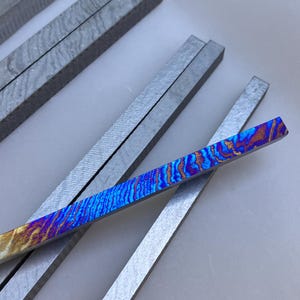 Könnte beinhalten: Nahaufnahme mehrerer rechteckiger Metallstäbe. Ein Stab weist ein lebendiges, farbenfrohes Muster aus Blau, Orange und Lila auf. Andere Stäbe sind einfarbig grau. Die Stäbe sind auf einer weißen Oberfläche angeordnet.