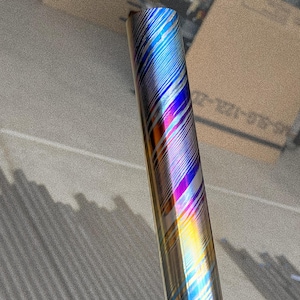 May include: A cylindrical object with a metallic finish, featuring a vibrant pattern of diagonal stripes in shades of blue, purple, orange, and silver. The surface reflects light, creating a rainbow effect. The object is set against a neutral background.