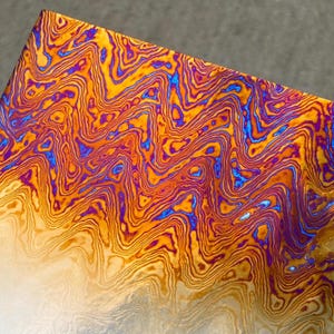 May include: Close-up of a metal surface with a vibrant, swirling pattern. The design features a blend of orange, yellow, blue, and purple hues, creating a dynamic and textured appearance. The metal has a polished, reflective quality.