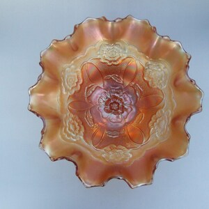 May include: An iridescent orange carnival glass bowl with a ruffled edge. The bowl features a floral pattern in the centre and around the sides. The glass has a glossy finish and reflects light, creating a shimmering effect.