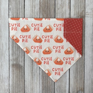 May include: A dog bandana with a repeating pattern of orange pie slices with smiling faces and the words "CUTIE PIE" in red. The bandana has a red polka dot accent on one corner. The background is a light wood grain.