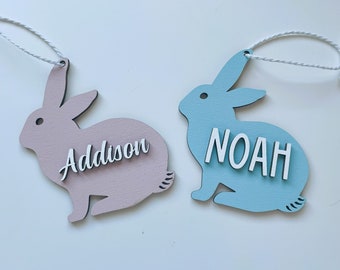May include: Two wooden bunny shaped ornaments with names Addison and Noah. The bunny with the name Addison is pink and the bunny with the name Noah is blue.