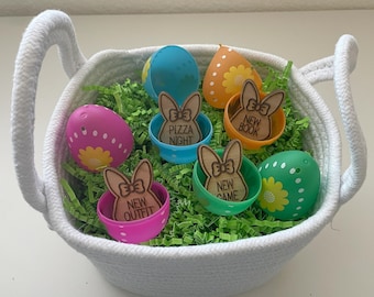 May include: A white wicker basket filled with green shredded paper and colorful plastic Easter eggs. Each egg has a wooden bunny cutout with a different message: "Pizza Night", "New Book", "New Game", and "New Outfit".