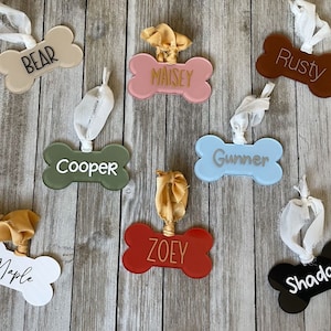 May include: Eight dog bone-shaped ornaments with different names written on them. The ornaments are made of different materials and colors, including white, black, red, blue, green, brown, and pink. The ornaments are hanging from a wooden surface.