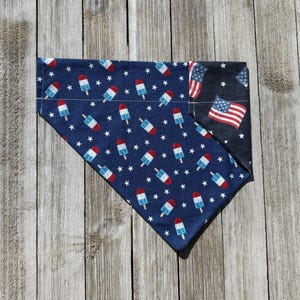 May include: A reversible dog bandana featuring a patriotic theme. One side has a navy blue background with white stars and red, white, and blue popsicle graphics. The other side displays the American flag on a dark blue background.