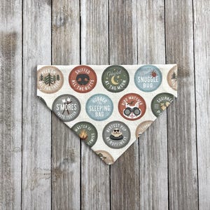 May include: A cream-colored dog bandana with a variety of circular designs featuring camping and outdoor-themed text and graphics. The bandana has a triangular shape and is made of fabric. The text includes phrases like "Snuggle Bug" and "S'mores Taste Tester."