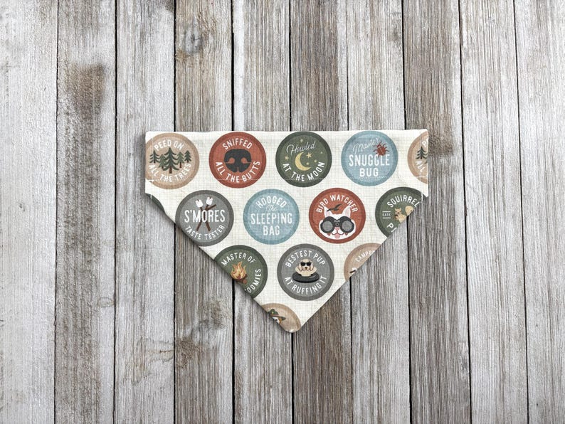 May include: A cream-colored dog bandana with a variety of circular designs featuring camping and outdoor-themed text and graphics. The bandana has a triangular shape and is made of fabric. The text includes phrases like "Snuggle Bug" and "S'mores Taste Tester."