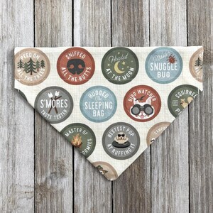 May include: A cream-colored dog bandana with various circular designs. Each circle features a different camping-themed illustration and text, such as "S'mores Taste Tester" and "Bestest Pup at Ruffing It". The bandana is triangular in shape.