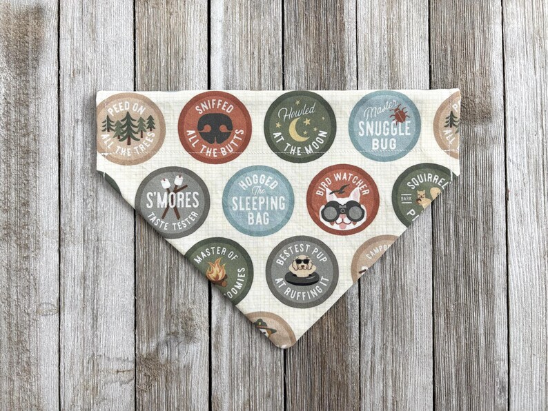 May include: A cream-colored dog bandana with various circular designs. Each circle features a different camping-themed illustration and text, such as "S'mores Taste Tester" and "Bestest Pup at Ruffing It". The bandana is triangular in shape.