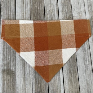 May include: A white and orange plaid dog bandana. The bandana is made of a soft, woven fabric and has a triangular shape.