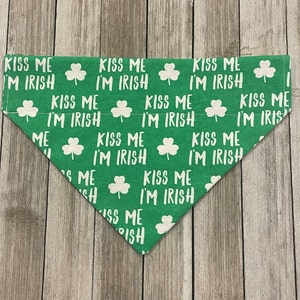 May include: Green dog bandana with white text and shamrock pattern. The text reads "KISS ME I'M IRISH" repeated across the fabric. The bandana is triangular in shape and made of fabric.