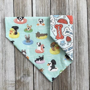 May include: A dog bandana with a light blue background featuring various dog breeds on inflatable floats. The reverse side has a white background with red and blue illustrations of bones, watermelon, and paw prints. The bandana is folded in a triangle shape.
