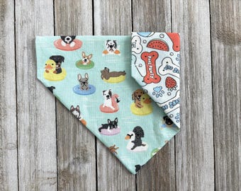 May include: A dog bandana with a light blue background featuring various dog breeds on inflatable floats. The reverse side has a white background with red and blue illustrations of bones, watermelon, and paw prints. The bandana is folded in a triangle shape.