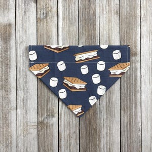 May include: A navy blue dog bandana with a repeating pattern of s'mores and marshmallows. The bandana is triangular in shape and made of fabric. The s'mores are brown and white, and the marshmallows are white.