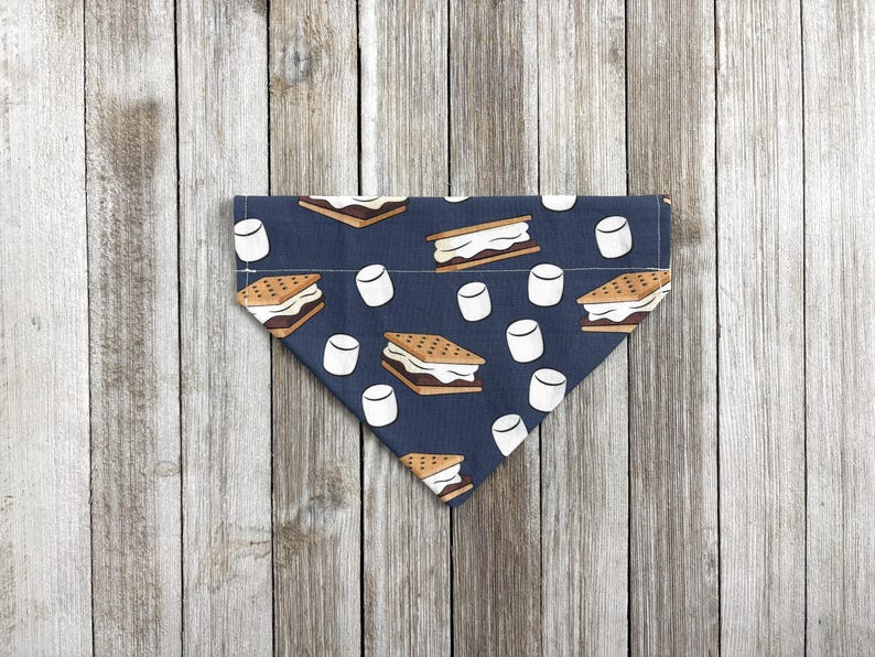 May include: A navy blue dog bandana with a repeating pattern of s'mores and marshmallows. The bandana is triangular in shape and made of fabric. The s'mores are brown and white, and the marshmallows are white.