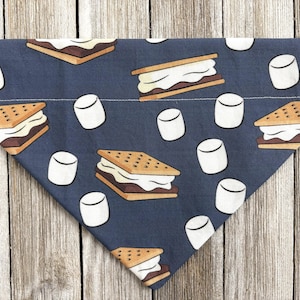 May include: A navy blue dog bandana with a repeating pattern of s'mores and marshmallows. The s'mores are brown, white, and tan, while the marshmallows are white. The bandana is triangular in shape.