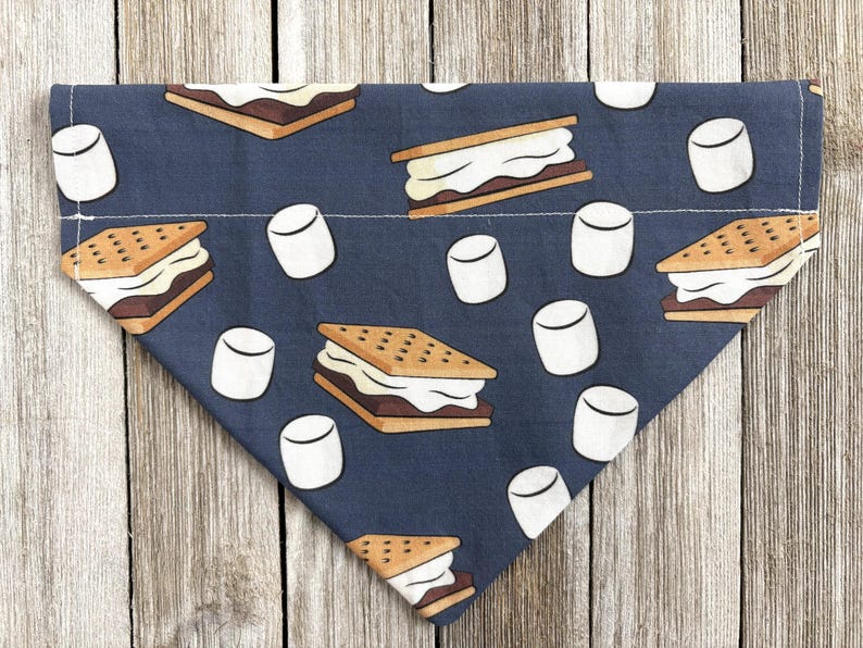 May include: A navy blue dog bandana with a repeating pattern of s'mores and marshmallows. The s'mores are brown, white, and tan, while the marshmallows are white. The bandana is triangular in shape.