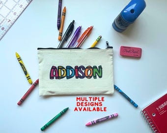 May include: A white canvas pencil case with a zipper closure. The name "ADDISON" is printed in colorful letters on the front of the case. The text "MULTIPLE DESIGNS AVAILABLE" is printed in red below the name.
