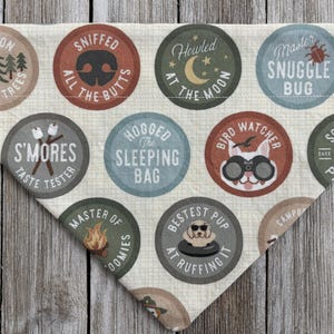 May include: A beige dog bandana with various circular badges. The badges feature phrases like "Sniffed All The Butts," "S'mores Taste Tester," and "Bestest Pup At Ruffing It." The bandana is made of a soft fabric and has a triangular shape.