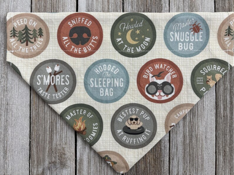 May include: A beige dog bandana with various circular badges. The badges feature phrases like "Sniffed All The Butts," "S'mores Taste Tester," and "Bestest Pup At Ruffing It." The bandana is made of a soft fabric and has a triangular shape.