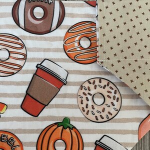 May include: A white fabric with brown stripes and a pattern of donuts, coffee cups, pumpkins, and footballs. The fabric has a brown and beige polka dot pattern on the edge.