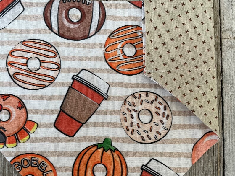 May include: A white fabric with brown stripes and a pattern of donuts, coffee cups, pumpkins, and footballs. The fabric has a brown and beige polka dot pattern on the edge.