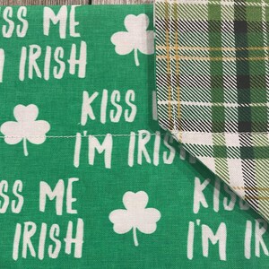 May include: Green fabric with the text "KISS ME I'M IRISH" in white, along with white shamrock designs. The corner features a green, black, and gold plaid pattern. A festive item for St. Patrick's Day.
