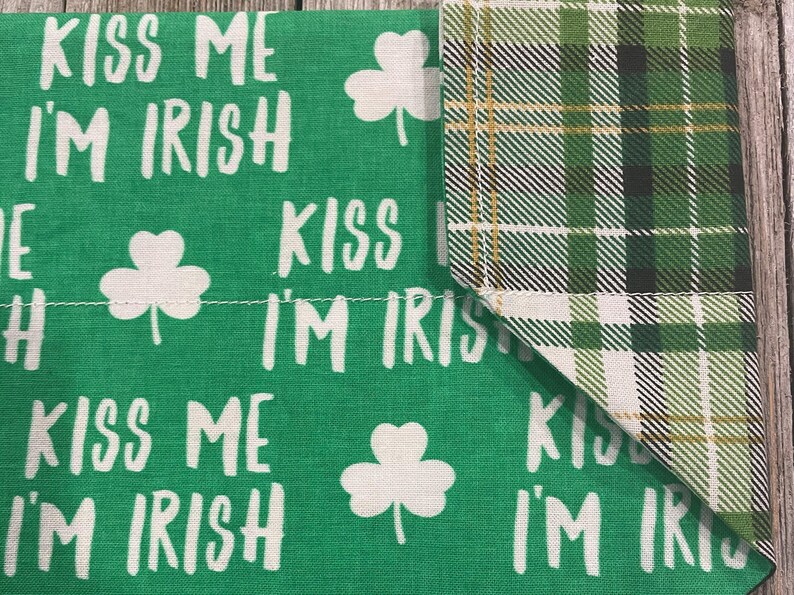 May include: Green fabric with the text "KISS ME I'M IRISH" in white, along with white shamrock designs. The corner features a green, black, and gold plaid pattern. A festive item for St. Patrick's Day.