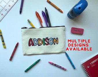 May include: A white canvas pencil case with a zipper closure. The case has the name "ADDISON" printed on it in colorful, block letters. The text "MULTIPLE DESIGNS AVAILABLE" is printed in red letters on a white background. The case is surrounded by crayons and pencils.