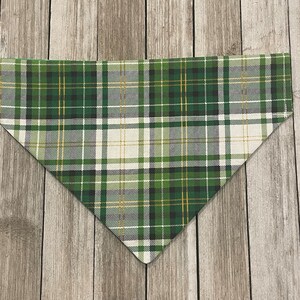 May include: Green, white, and gold plaid dog bandana. The bandana is triangular in shape and features a classic plaid pattern with varying shades of green, white, and gold. The fabric is folded and ready to be worn.