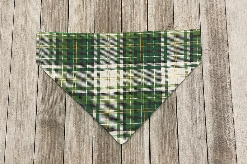 May include: Green, white, and gold plaid dog bandana. The bandana is triangular in shape and features a classic plaid pattern with varying shades of green, white, and gold. The fabric is folded and ready to be worn.