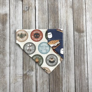 May include: A dog bandana with a camping theme. One side features circular badges with text like "S'mores Taste Tester" and "Sniffed All The Butts." The other side has a dark blue pattern with marshmallows and a s'more.