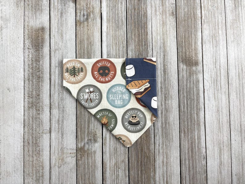 May include: A dog bandana with a camping theme. One side features circular badges with text like "S'mores Taste Tester" and "Sniffed All The Butts." The other side has a dark blue pattern with marshmallows and a s'more.