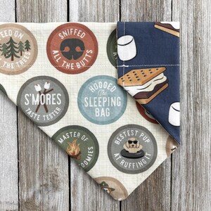 May include: A dog bandana with a camping theme. The bandana features various circular designs with text like "S'mores Taste Tester" and "Bestest Pup at Ruffing It." One side has a blue background with marshmallow and s'more graphics.