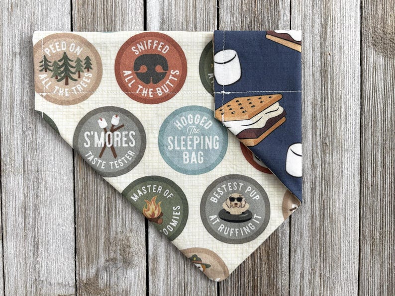 May include: A dog bandana with a camping theme. The bandana features various circular designs with text like "S'mores Taste Tester" and "Bestest Pup at Ruffing It." One side has a blue background with marshmallow and s'more graphics.
