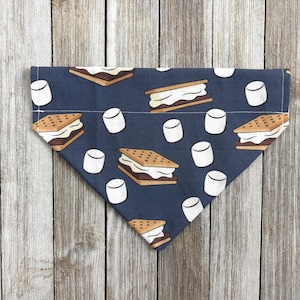 May include: A navy blue dog bandana featuring a repeating pattern of s'mores and marshmallows. The s'mores are brown and white, and the marshmallows are white. The bandana is folded in a triangle shape.