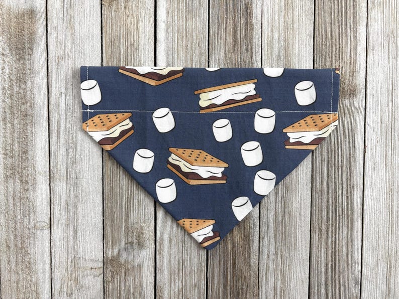 May include: A navy blue dog bandana featuring a repeating pattern of s'mores and marshmallows. The s'mores are brown and white, and the marshmallows are white. The bandana is folded in a triangle shape.