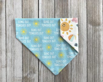 May include: A blue dog bandana with the text "SUNS OUT TONGUES OUT" and yellow sun graphics. The reverse side features a smiling sun, beach ball, and floral pattern. The bandana is folded on a wooden surface.