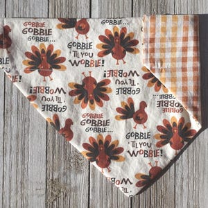May include: A triangular pet bandana featuring a cream-colored fabric with a repeating pattern of brown turkeys and the text GOBBLE GOBBLE GOBBLE... GOBBLE 'TIL YOU WOBBLE! The reverse side of the bandana displays a classic orange and white gingham pattern.